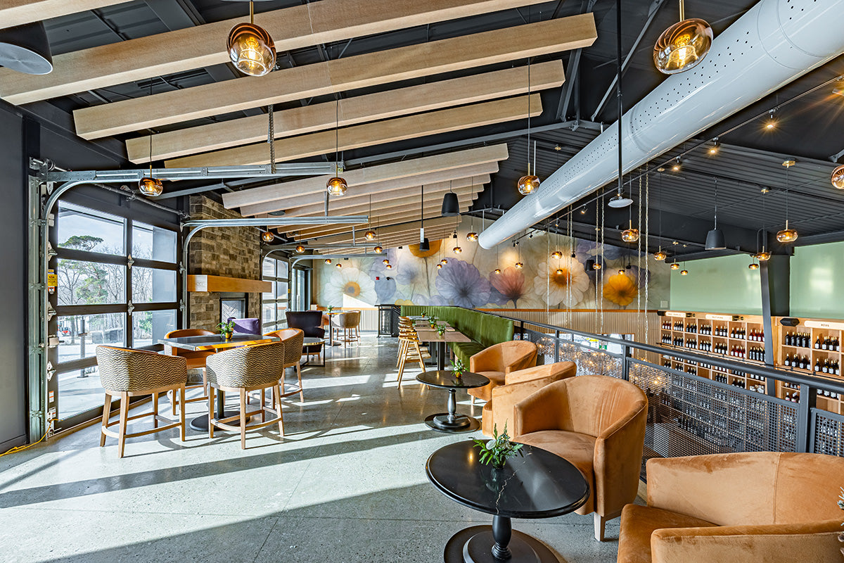 Skye Chase's modern mezzanine interior with exposed ceiling beams, hanging pendant lights, upholstered bar stools, round tables, and a glass railing overlooking shelves of bottles.