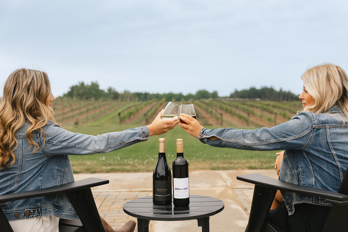 Two women in denim jackets clinking wine glasses at an outdoor table with two wine bottles, set against the Skye Chase vineyard landscape.