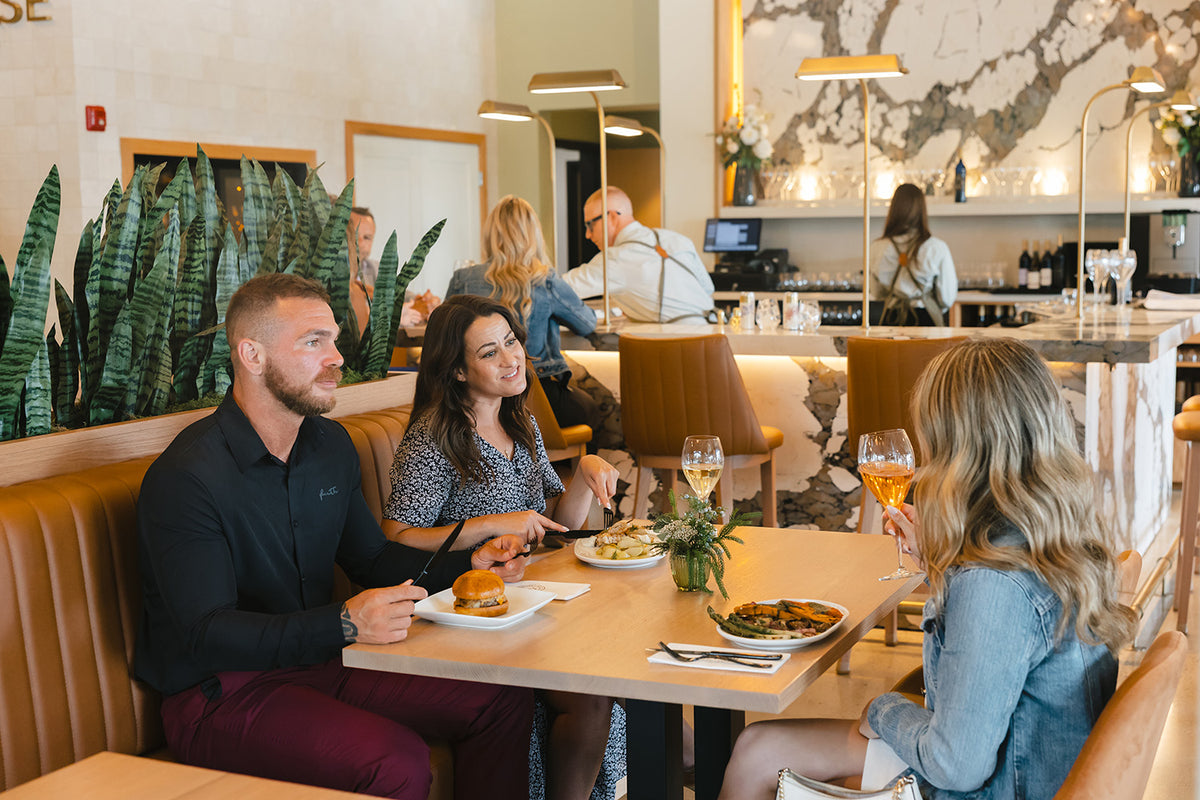 A group of friends dining together at a booth, with plates of food and wine glasses on the table in a warmly lit interior setting.