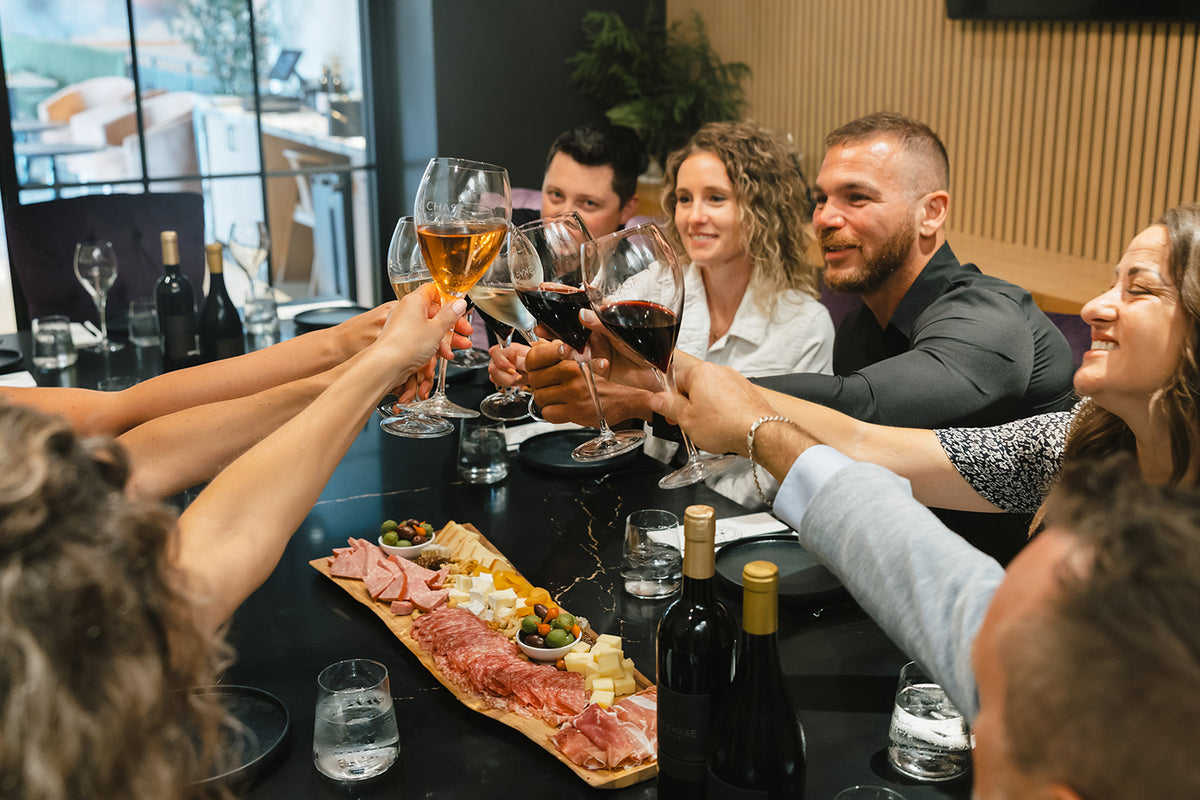 A group of friends and family raising wine glasses in a toast on a dark dining table, with bottles of wine, a charcuterie board, and a modern restaurant interior in the background.