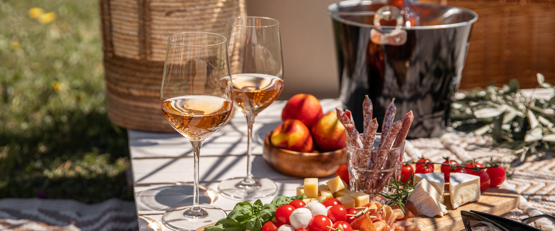 Table outdoors in the sun with wine glasses, food, ice bucket, picnic basket.